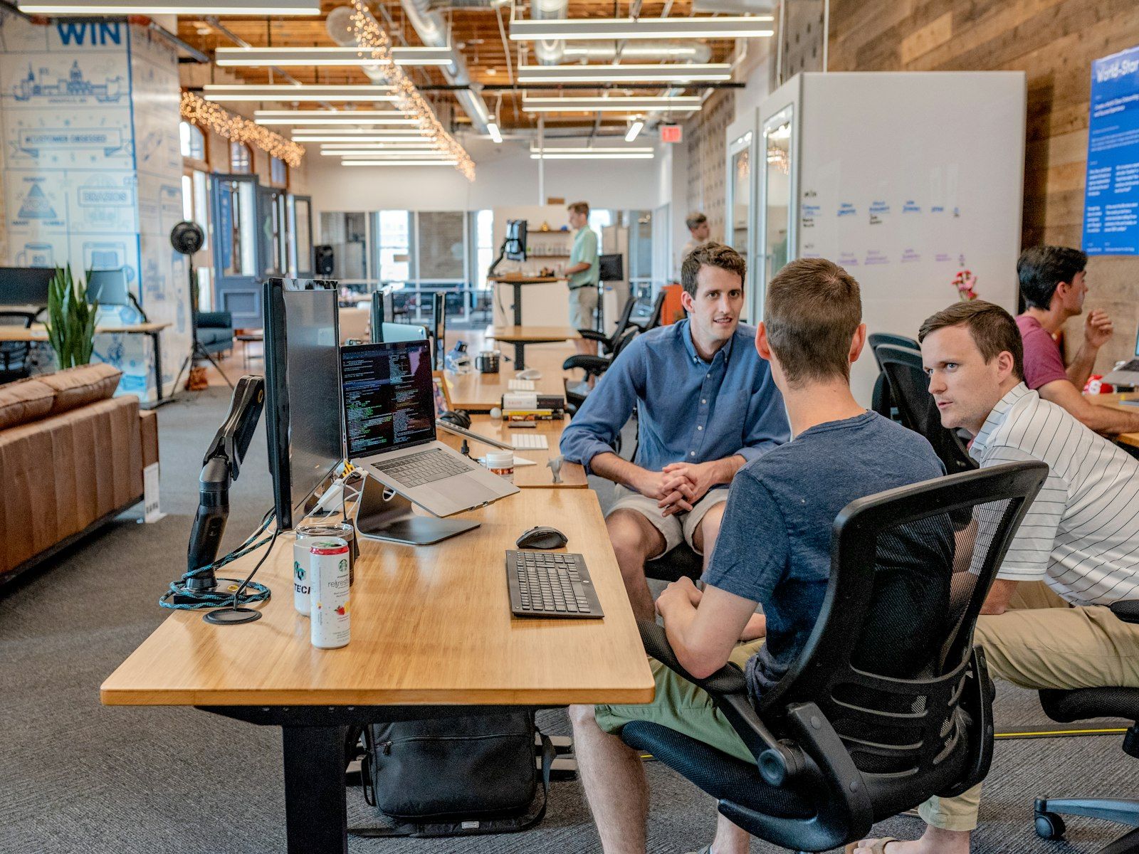 Three colleagues talking at a desk in an open-plan office, beside a monitor full of code