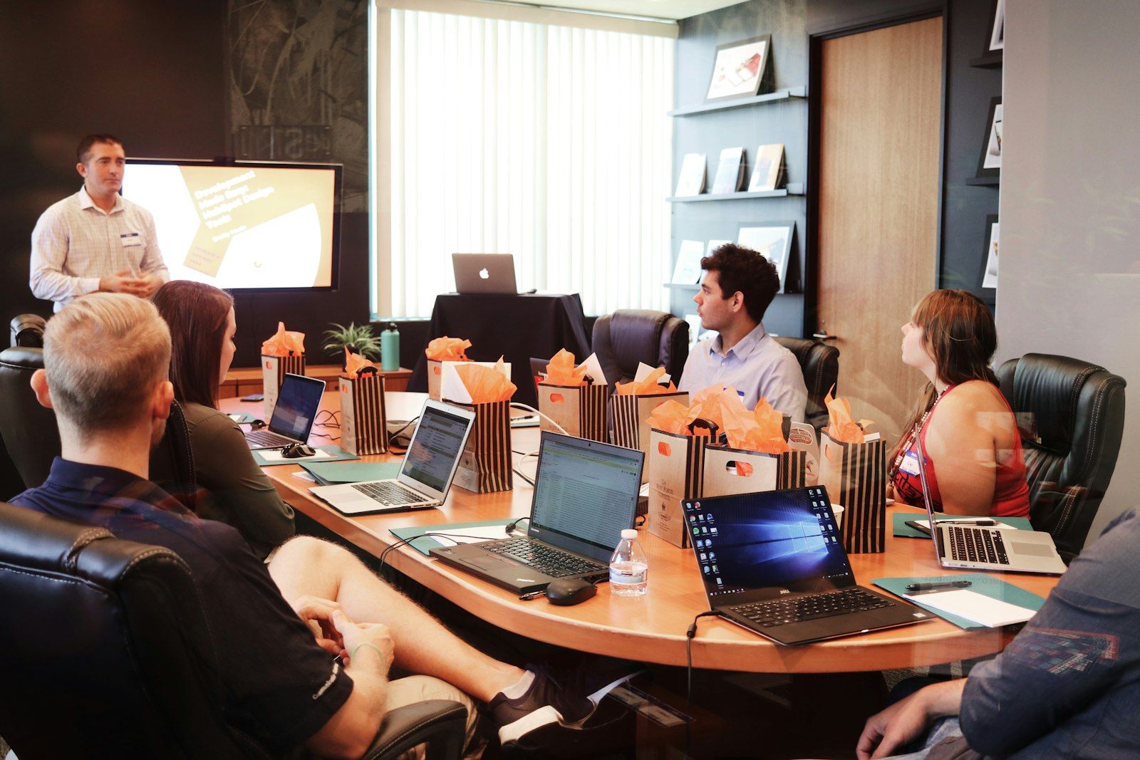A consultant presenting to five colleagues seated around a boardroom table with laptops open